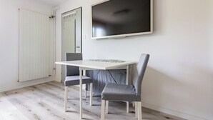 Una habitación con una mesa blanca y dos sillas grises, un televisor de pantalla plana grande montado en la pared y un radiador blanco.