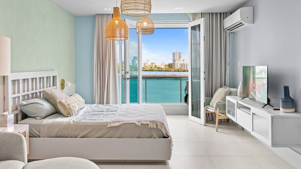 A modern bedroom with a bed, a desk, and a TV. It has a large window with a view of the city and a balcony. - Perfect Couples Getaway with Ocean View Location (San Juan)