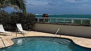 A clear blue pool with a curved edge, surrounded by a low wall and a railing. There are two empty lounge chairs and a palm tree in the background. - Perfect Couples Getaway with Ocean View Location (San Juan)