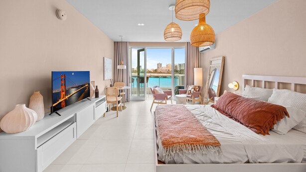 A modern hotel room with a bed, a TV, a desk, and a view of a cityscape and water. - Stylish 1BR with Balcony & Prime Location (San Juan)