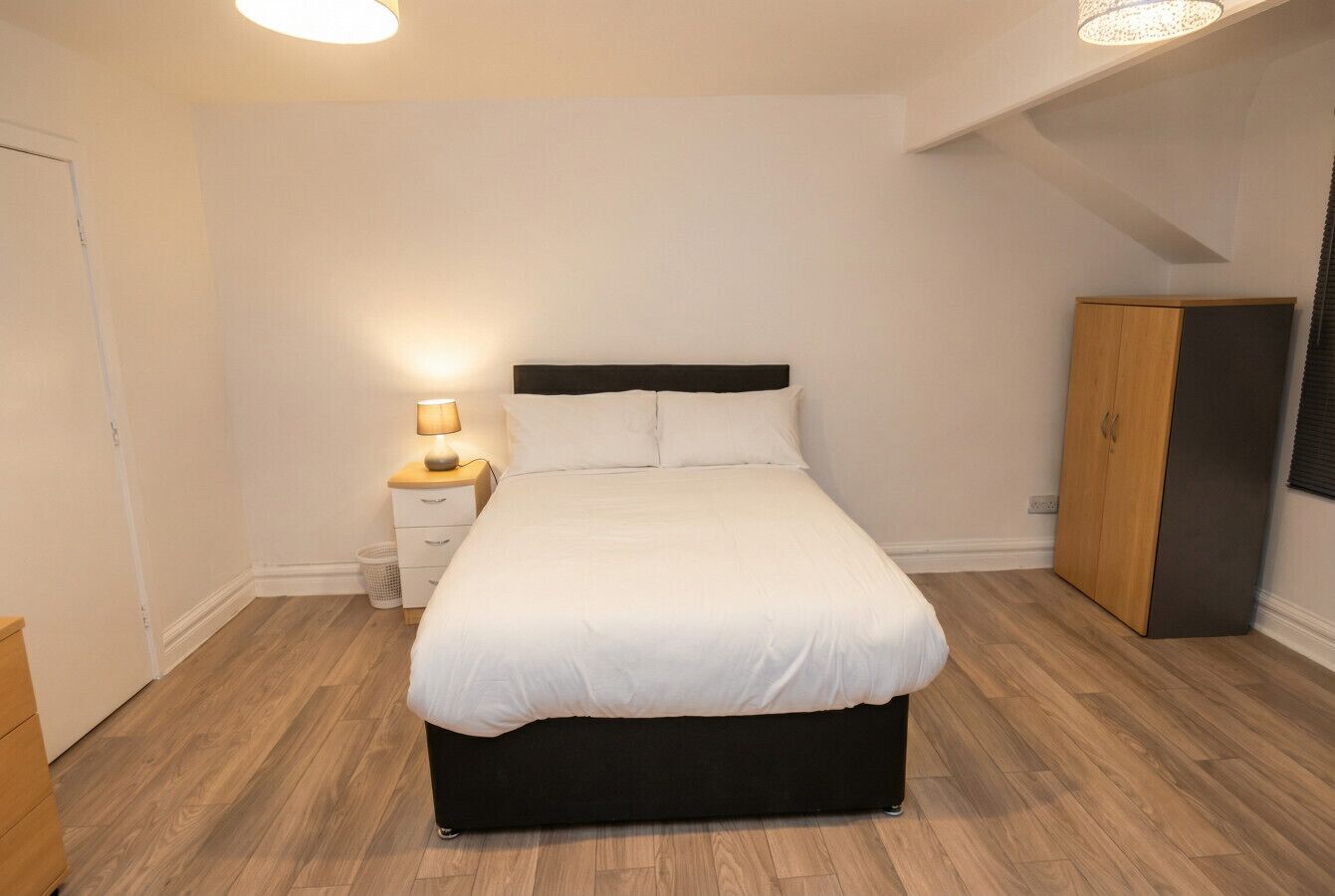 A bedroom with a bed, bedside table, lamp, wardrobe, and a wooden floor.