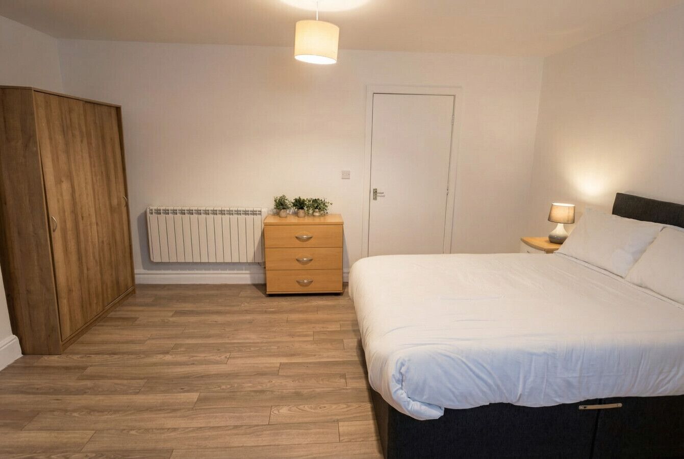 Ein Schlafzimmer mit einem Bett, einem Holzschrank, einem Nachttisch mit Lampe, einem Heizkörper und einer Tür.