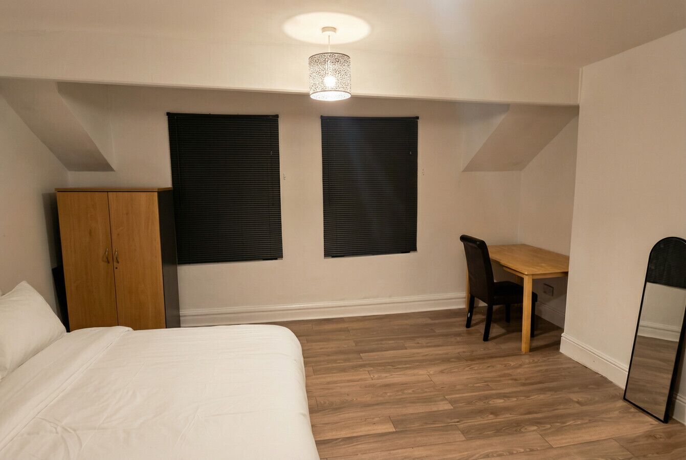 Ein Schlafzimmer mit Bett, einem Holzschrank, einem Esstisch mit Stühlen und zwei Fenstern mit Jalousien.