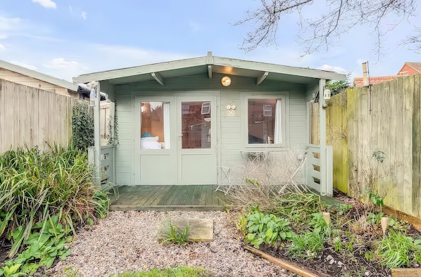 A small, light green shed with two windows and a door, surrounded by gravel and plants. - Bay Tree Cottage (King's Lynn)