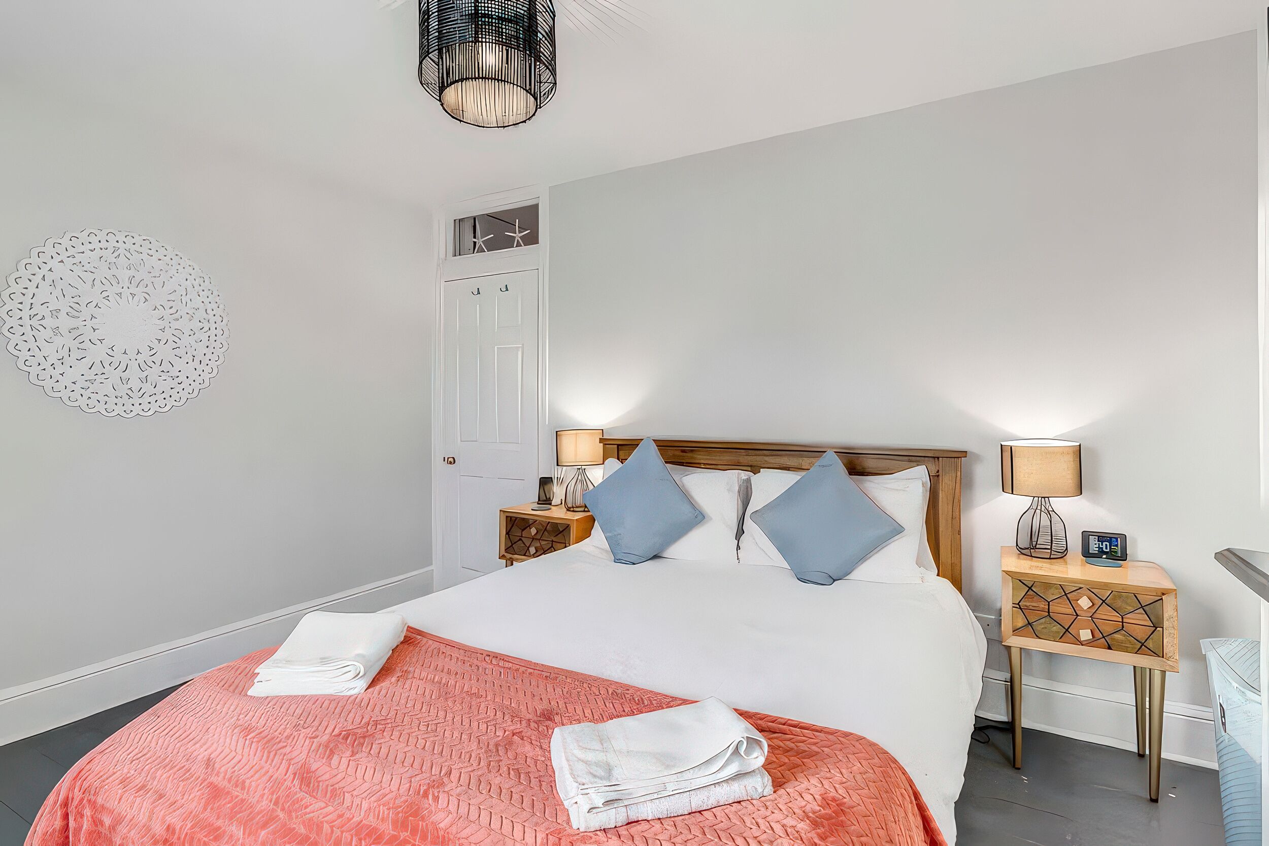 Een net opgemaakt bed met een roze sprei, twee blauwe kussens en opgevouwen witte handdoeken. Er zijn twee nachtkastjes met lampen, een houten hoofdbord en een rond decoratie aan de muur.