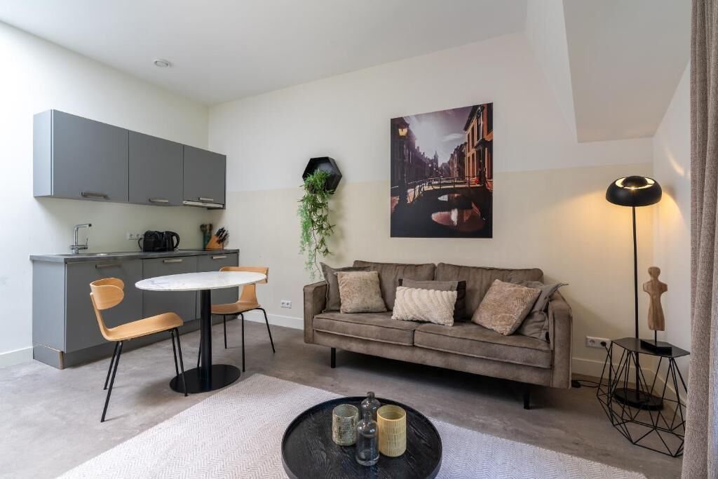 Una sala de estar moderna con un sofá gris, una mesa redonda y una mesa de centro. Cuenta con una zona de cocina con armarios grises y una planta en la pared.