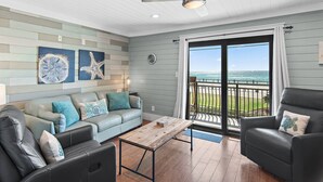 Ein Wohnzimmer mit hellblauer Wand, einem Holztisch, einem Sofa mit blauen Kissen und einem schwarzen Sessel. Ein großes Fenster bietet Blick auf das Meer.