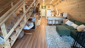 A cozy cabin interior with a wooden staircase, a green sofa, a kitchen area, and a dining table. - A Frame Cabin In The Woods (Omaha)