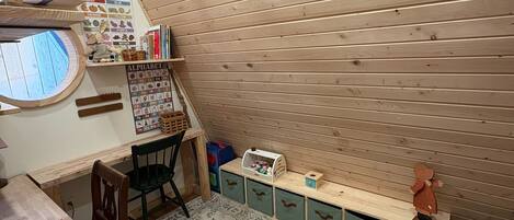 Una habitación acogedora con techo de listones de madera, un ventanal redondo, un escritorio de madera, una silla verde, un tapete estampado y cajones para guardar objetos.