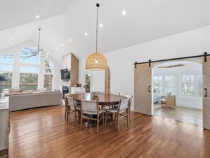 A spacious dining area with a wooden table and chairs, a large woven pendant light, and a view of the outdoors through large windows. - Tree House Escape - expansive decks and beautiful views! (Arley)