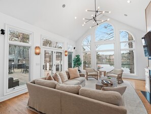 A spacious living area with a large beige sectional sofa, a coffee table, and wicker chairs. There are large windows offering views of a deck and outdoor scenery. - Tree House Escape - expansive decks and beautiful views! (Arley)