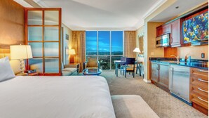 A hotel room with a large bed, a balcony with a city view, a kitchenette, and a dining area.