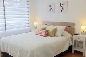 Un lit bien fait, avec une couette blanche et deux oreillers, une table de chevet avec une lampe et une fenêtre avec des stores.
