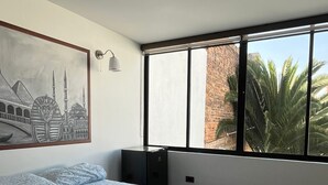 A bedroom with a large bed, a painting of a mosque, a window with a view of palm trees, and a brick wall outside. - THE LONDON INN (Bogotá)