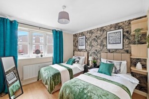 Ein Schlafzimmer mit zwei Betten, türkisfarbenen Vorhängen, einem Heizkörper und gerahmten Bildern an der Wand.