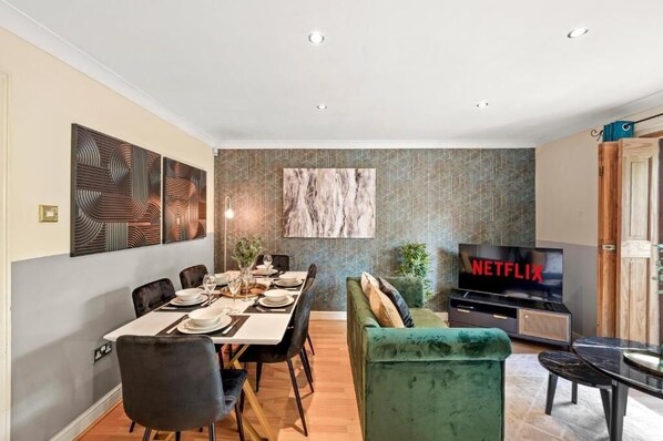 Ein modernes Wohnzimmer mit Essbereich, einem grünen Sofa, einem Fernseher, der 'NETFLIX' zeigt, und einer Wand mit Muster.