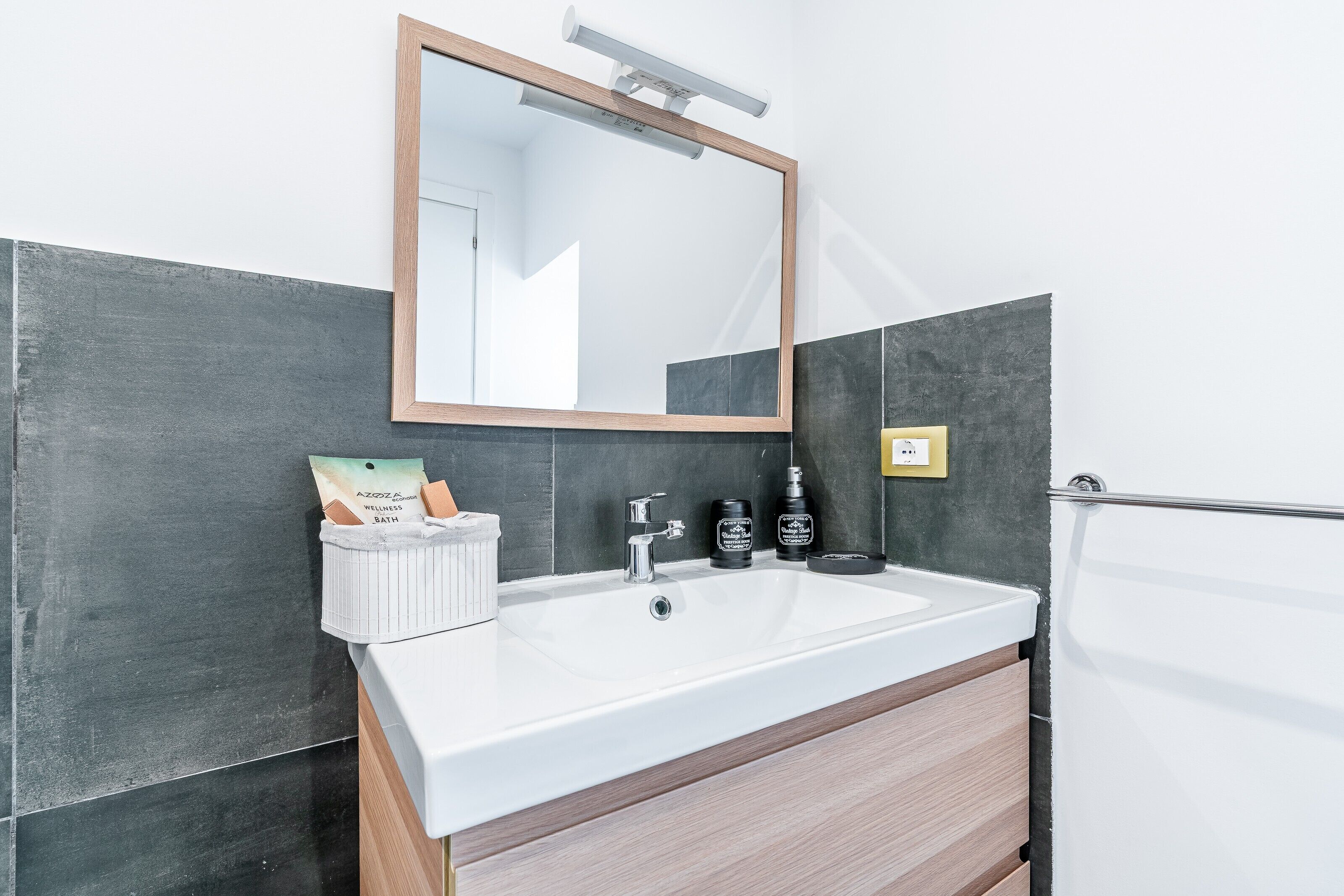 A bathroom with a white sink, dark tiled walls, a wooden cabinet, a mirror, and toiletries.