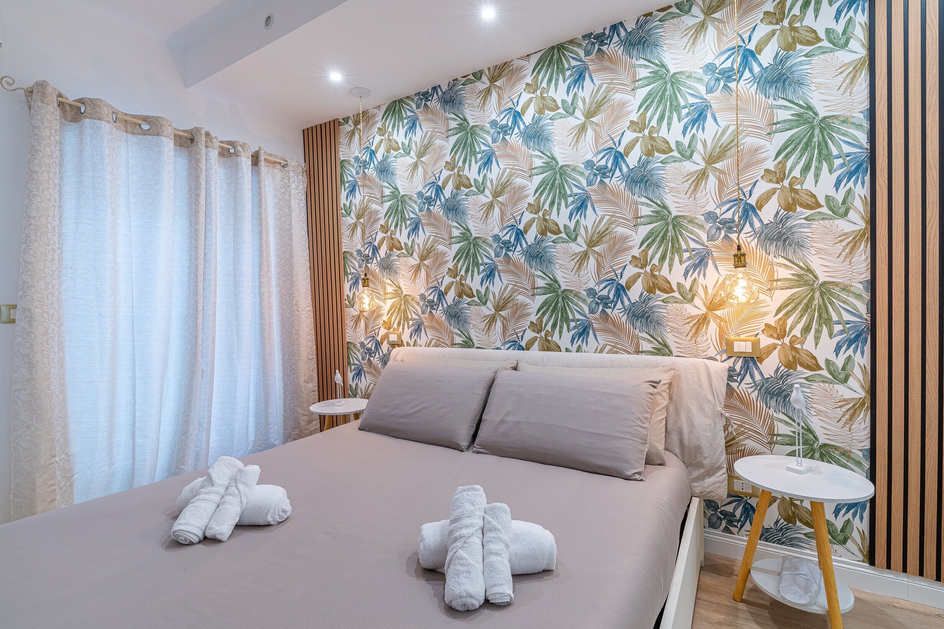 A bedroom with a bed, towels, a nightstand, a wall with a leaf pattern, and a wooden panel.