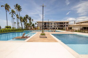 Área de piscina em resort, com várias piscinas, palmeiras e um edifício alto ao fundo.
