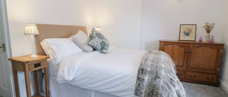 A neatly made bed with white linens and a grey throw blanket, a bedside table with a lamp, and a wooden cabinet with decorative items.