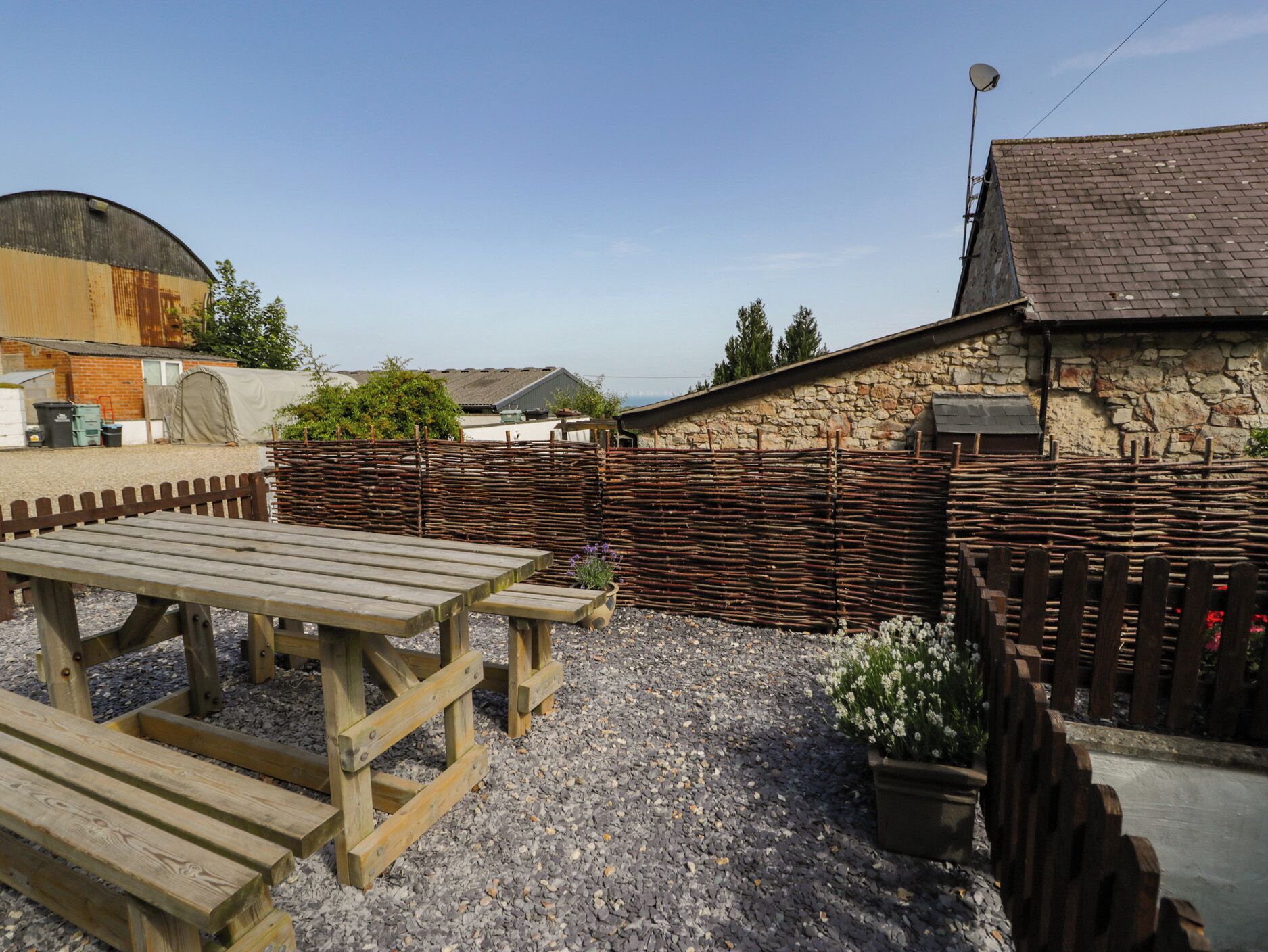 A rustic outdoor area with a wooden picnic table and benches, surrounded by a stone wall and a wooden fence.