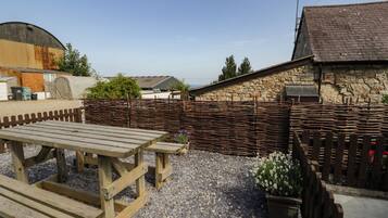 A rustic outdoor area with a wooden picnic table and benches, surrounded by a stone wall and a wooden fence.