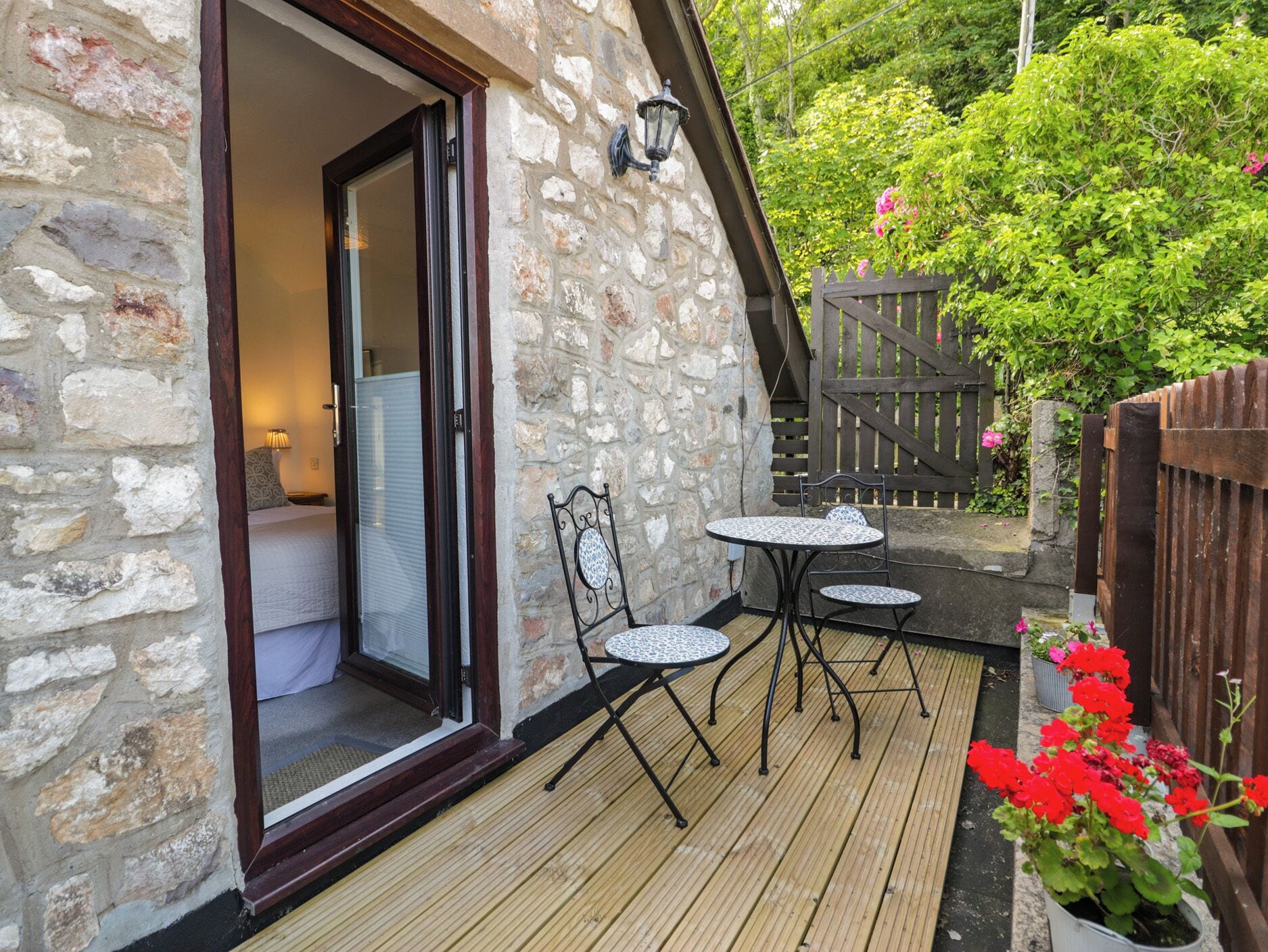 A stone building with a wooden deck, a glass door entrance, a small table with two chairs, and a wooden fence with greenery.