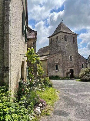 Un bâtiment historique en pierre, doté d’une tour centrale, d’une allée gravillonnée et entouré de verdure.