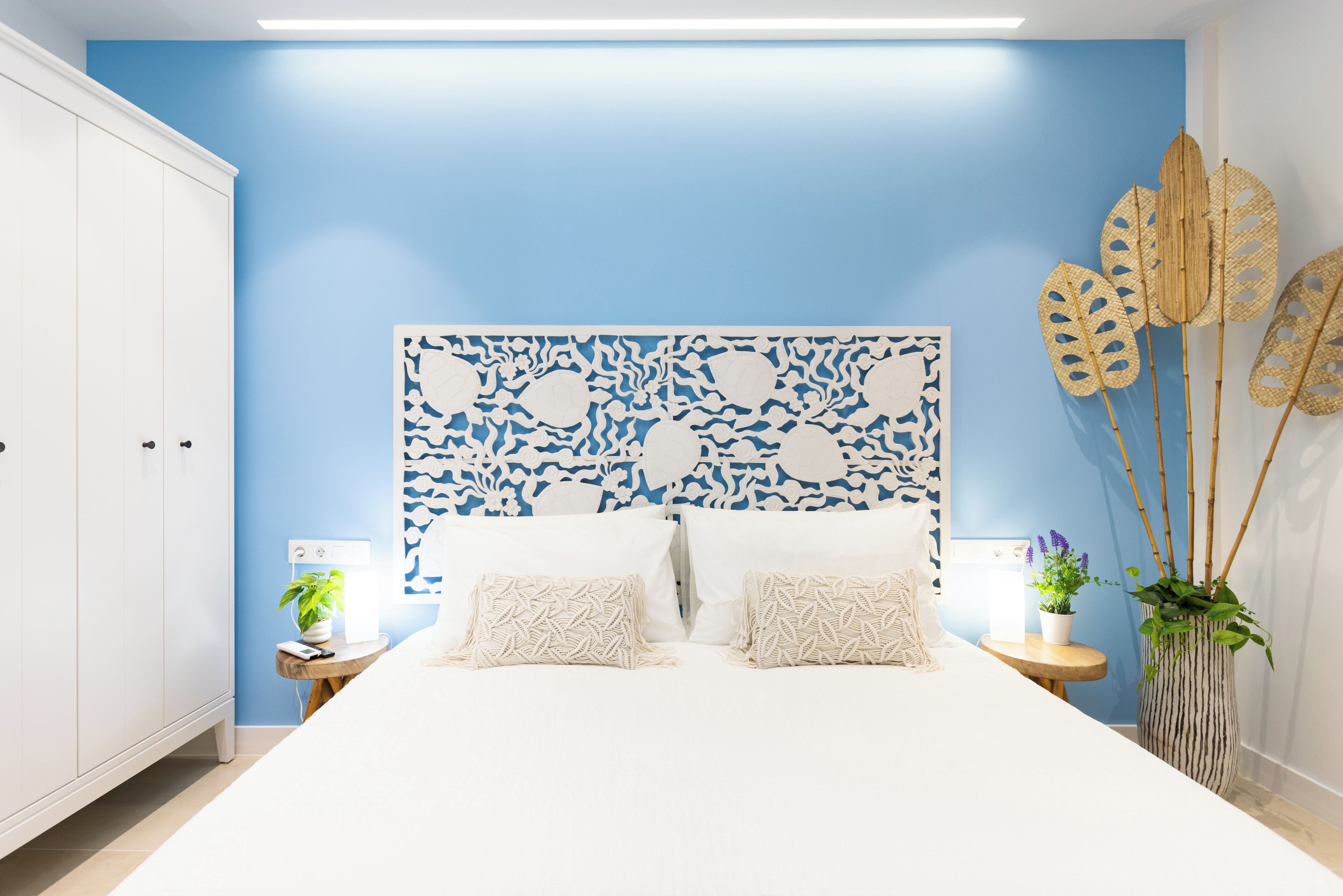 Ein Schlafzimmer mit einem weißen Bett, einer blauen Akzentwand, einem dekorativen Kopfteil, Nachttischen mit Lampen und einer Blumentopf-Pflanze.