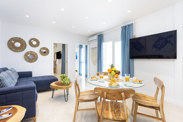 A modern living room with a blue sofa, wooden dining table with chairs, and wall-mounted decor.