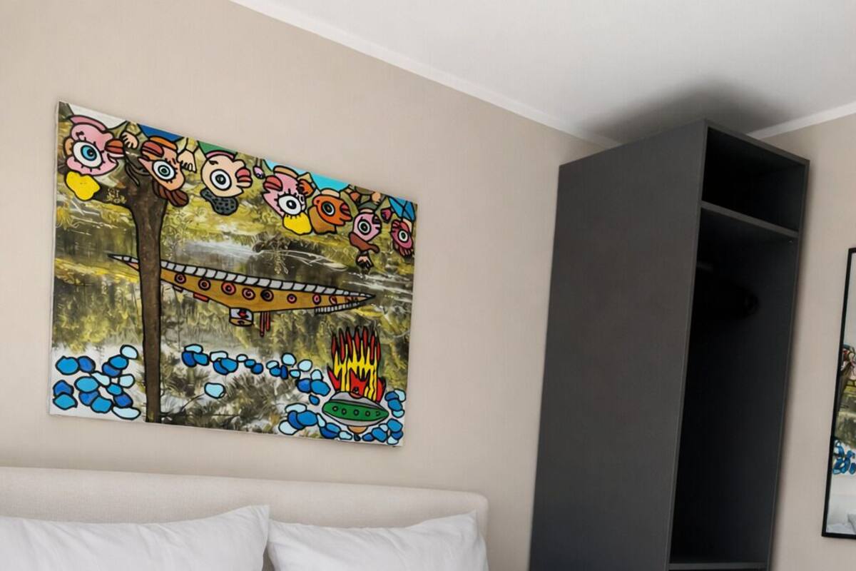 A hotel room with a bed, pillows, a gray blanket, and a wall with a colorful painting.