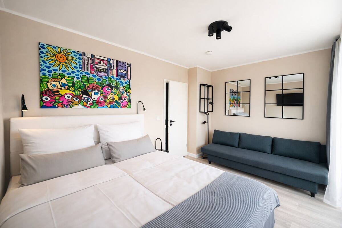 A modern bedroom with a bed, a sofa, a colorful artwork, and a ceiling light.