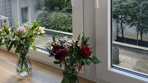 Et vindue med to vaser blomster på en træhylde, der viser udsigt over et boligområde med bygninger og træer.