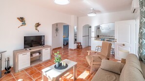 Ein modernes Wohnzimmer mit Fernseher, Sofa und Esstisch. Im Hintergrund ist eine Küche mit Mikrowelle und Schränken zu sehen.
