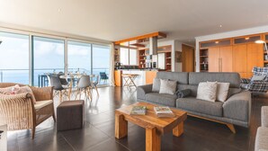 Ein großzügiger Wohnbereich mit grauem Sofa, Holz-Couchtisch, Korbmöbeln und Meerblick durch große Fenster.