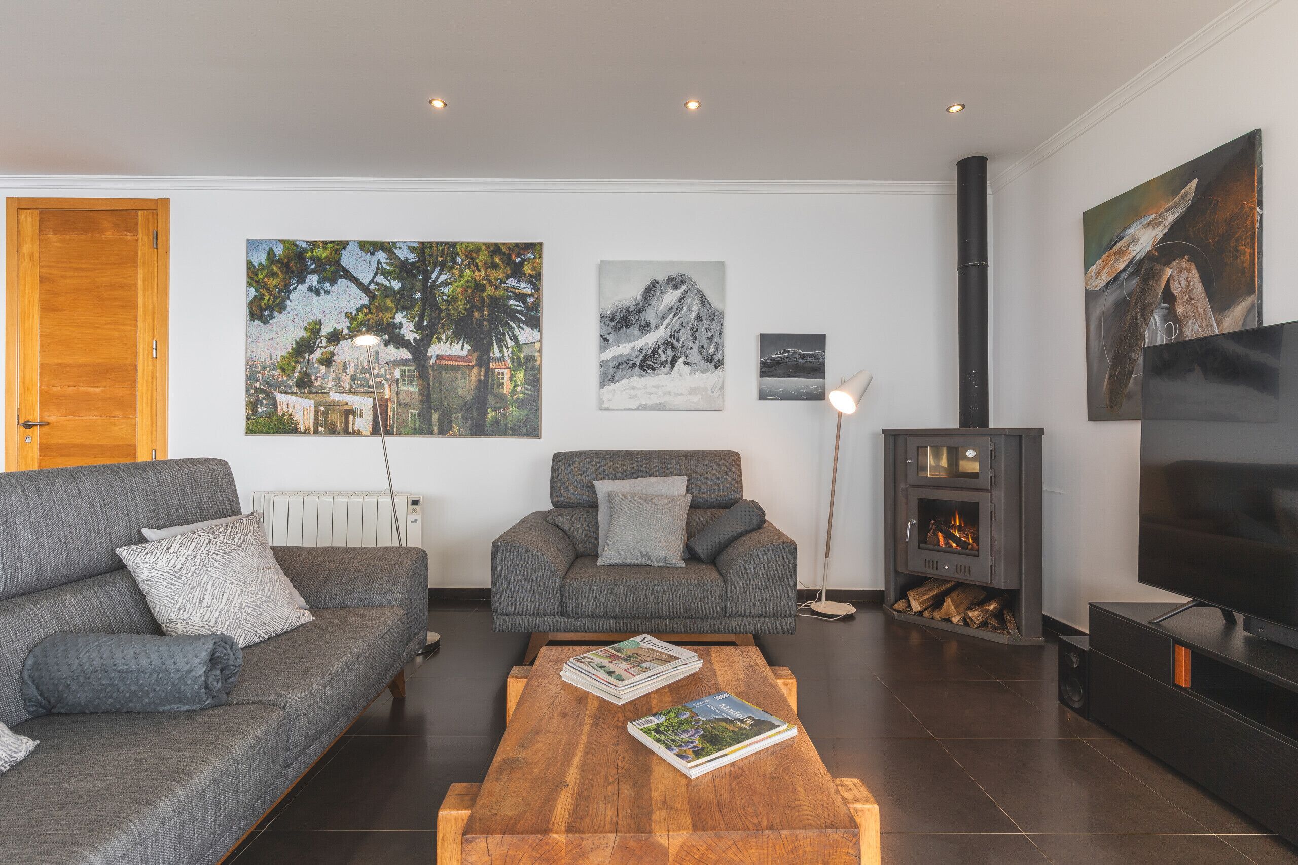 A modern living room with a grey sofa, a wooden coffee table with magazines, a grey armchair, a fireplace, and a TV.
