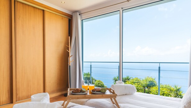 Ein Schlafzimmer mit großem Fenster mit Meerblick, einem hölzernen Nachttisch mit Frühstücksgebäck und einem ordentlich bezogenen Bett.