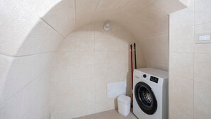 Une petite buanderie comprenant un lave-linge, une petite poubelle et un balai appuyé contre le mur.