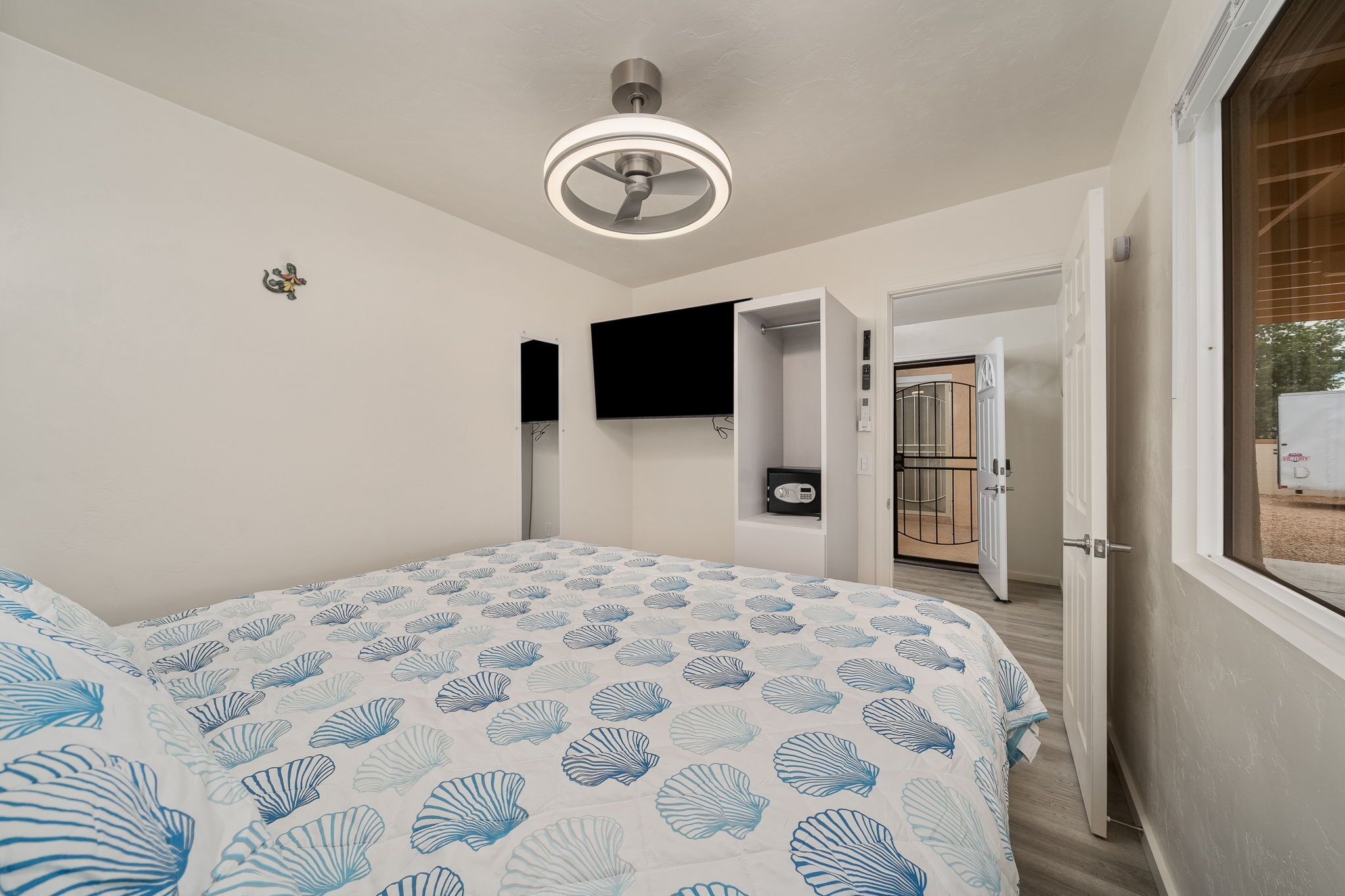 Une chambre à coucher comprenant un lit, un ventilateur de plafond, un téléviseur fixé au mur et une porte donnant sur une autre pièce.