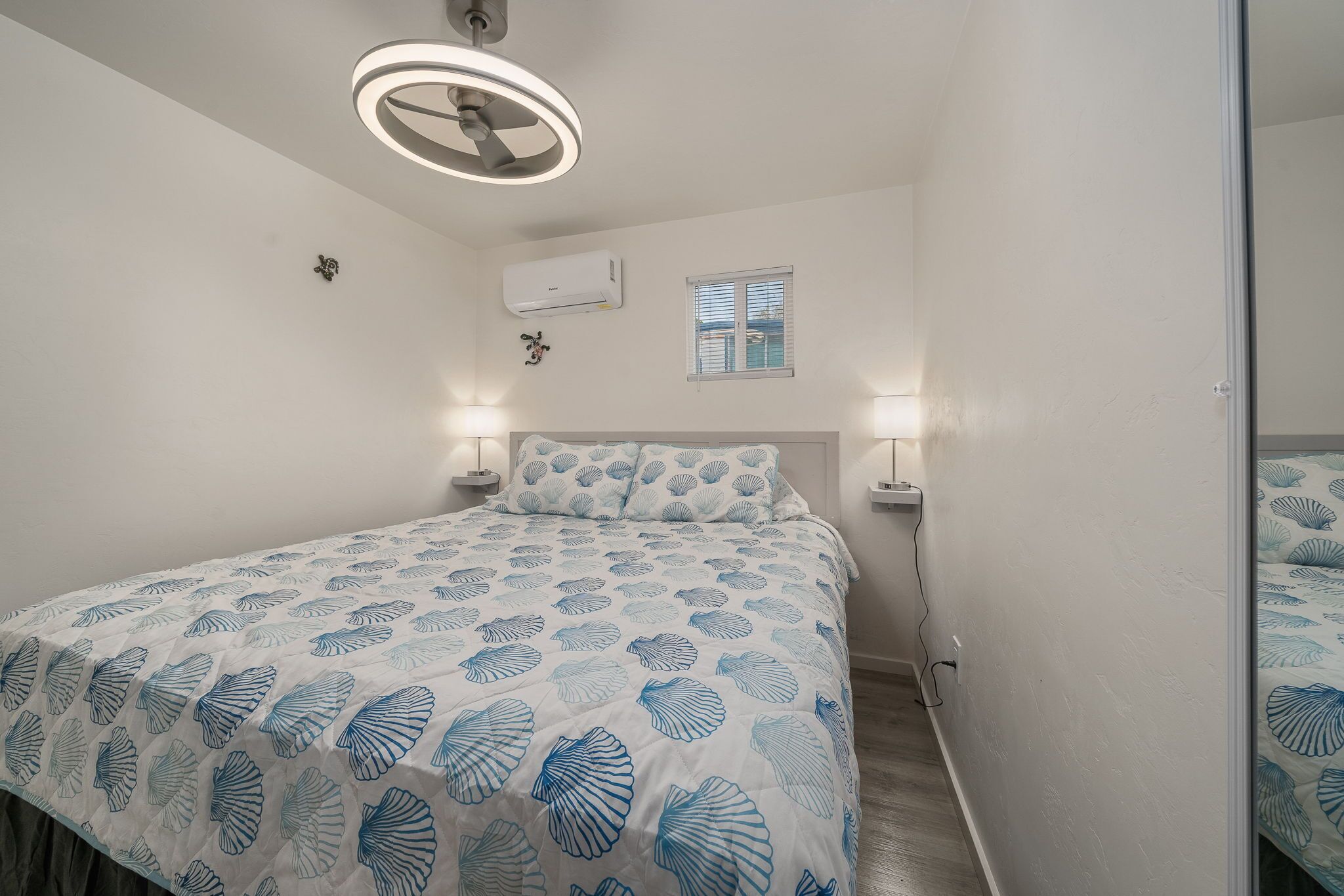 A bedroom with a bed, two bedside lamps, a ceiling fan, an air conditioning unit, and a window.