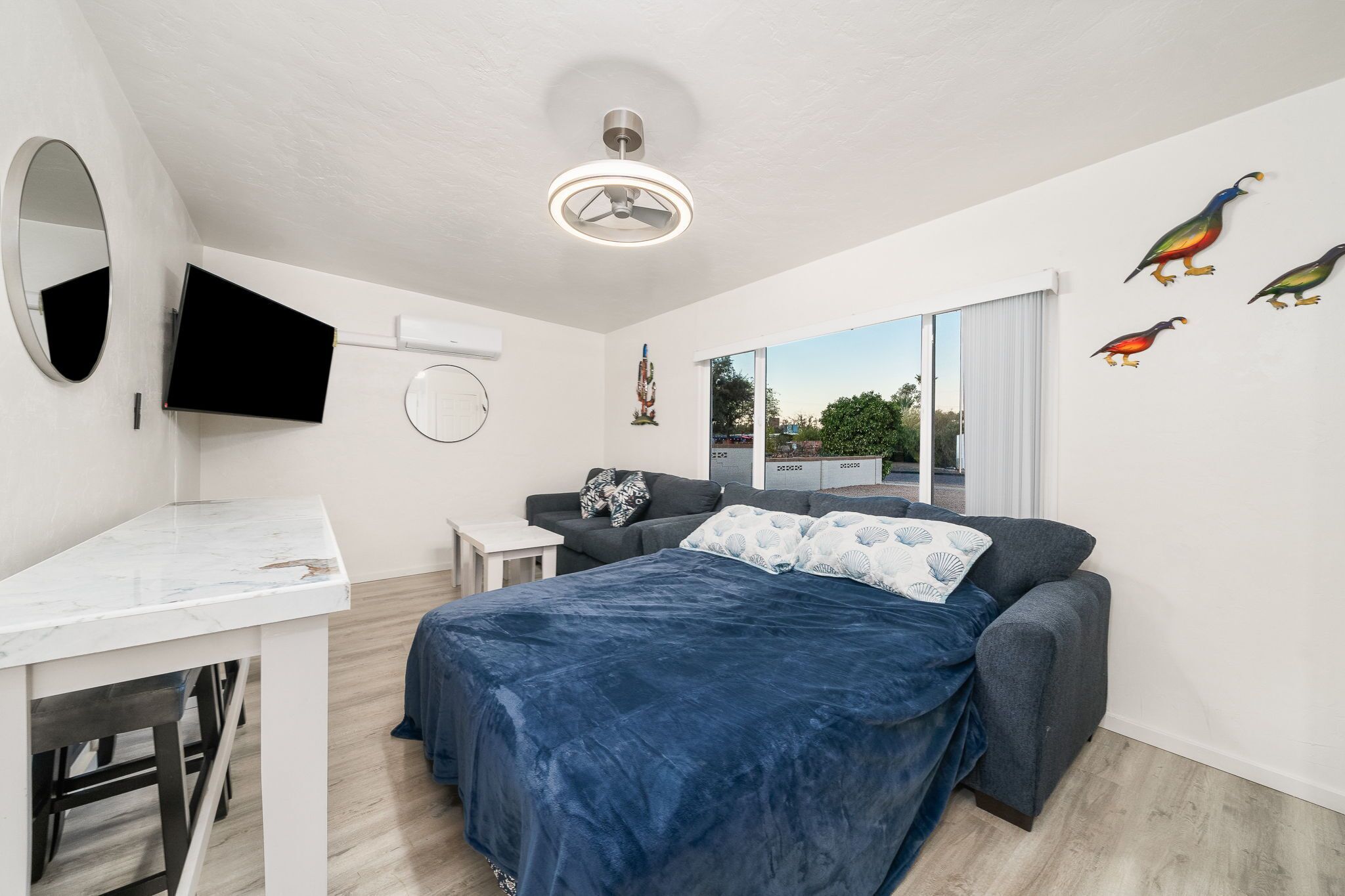 A bedroom with a bed, a desk, a chair, a TV, a ceiling fan, and a window with a view.