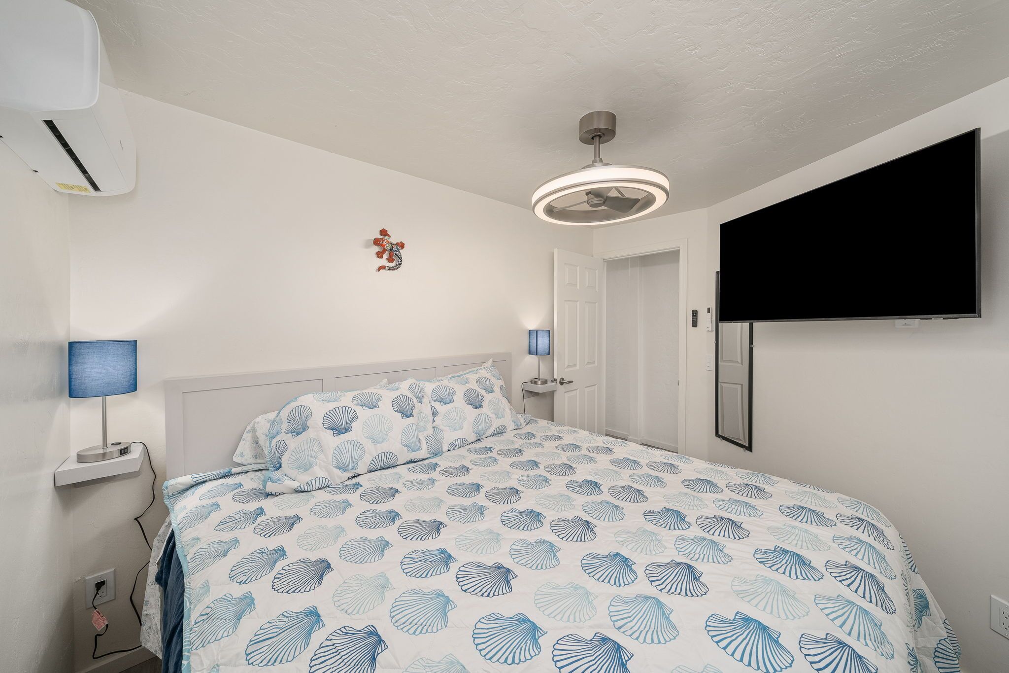 A bedroom with a bed, bedside lamps, a ceiling fan, a TV, and a wall-mounted air conditioner.