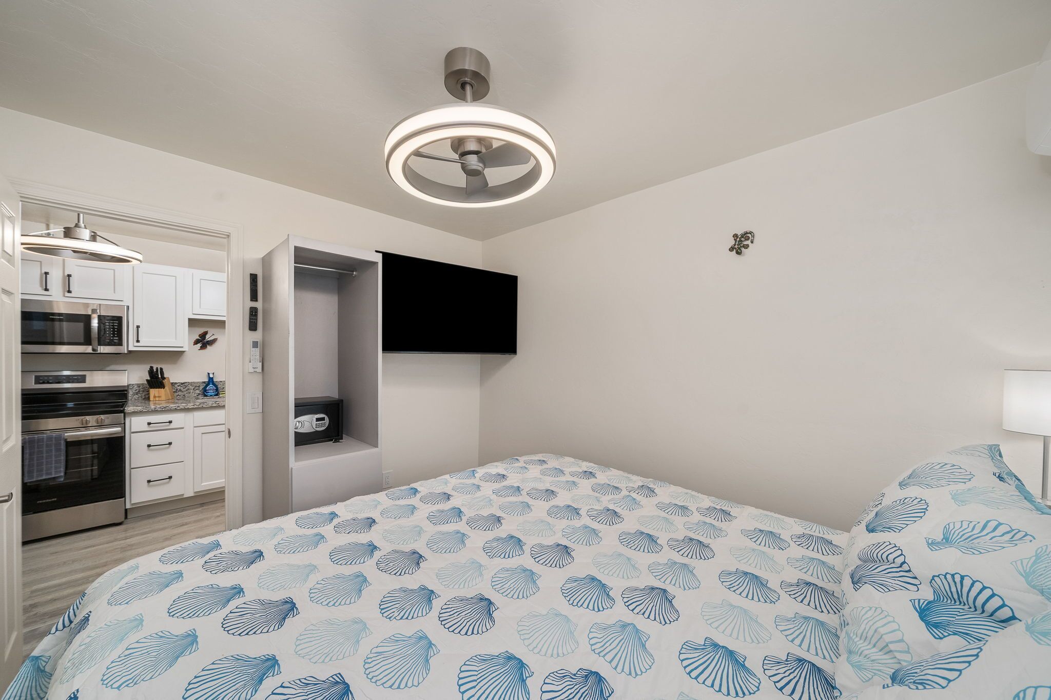 Een slaapkamer met een bed, een plafondventilator, een aan de muur bevestigde televisie en een keuken met kasten en apparatuur.