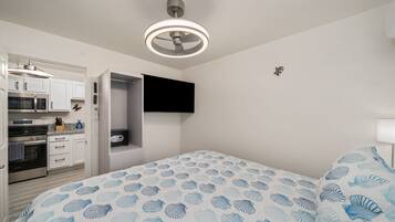 Een slaapkamer met een bed, een plafondventilator, een aan de muur bevestigde televisie en een keuken met kasten en apparatuur.
