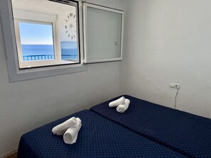 Ein Zimmer mit zwei Betten, einem Fenster mit Meerblick, einer Wanduhr und einer Steckdose.