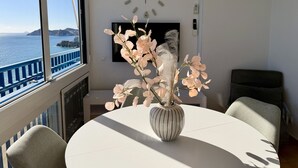 Une pièce avec une table ronde, des chaises, un vase de fleurs, une horloge murale et une vue sur la mer par la fenêtre.