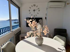 Ein Zimmer mit rundem Tisch, Stühlen, einer Vase mit Blumen, einer Wanduhr und Blick auf das Meer durch ein Fenster.