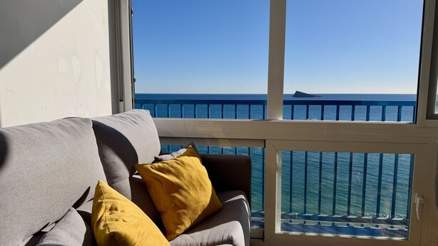 Une pièce avec un canapé gris, des coussins jaunes et une vue sur la mer, visible à travers la rambarde d’un balcon.