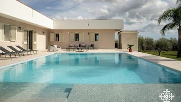 Piscina con sillas de descanso y una mesa, rodeada por un edificio con contraventanas y una palmera.