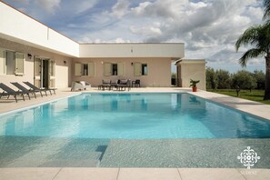 Une piscine entourée de chaises longues et d’une table, le tout bordé d’un bâtiment aux volets fermés et d’un palmier.
